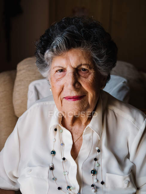 Ritratto di donna anziana felice in camicia bianca e con perline sul collo guardando la macchina fotografica a casa — Foto stock