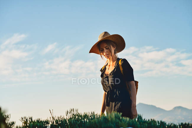 Turista femenina con sombrero de paja y mochila de pie en la naturaleza - foto de stock