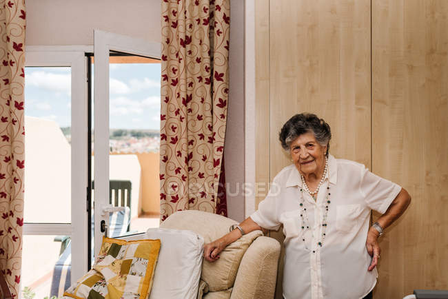 Старша жінка в білій сорочці з рукою на талії стоячи, спираючись на крісло вдома і дивлячись на камеру — стокове фото