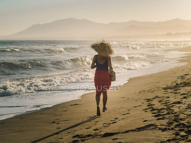 Back view of slim woman in hat and pareo running along coastline in bright sunlight — Stock Photo