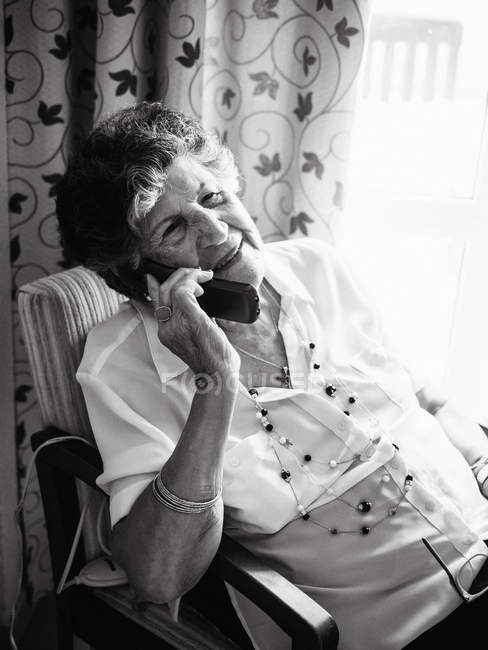 Feliz mujer mayor sonriendo y hablando en el teléfono móvil mientras mira hacia fuera en casa - foto de stock