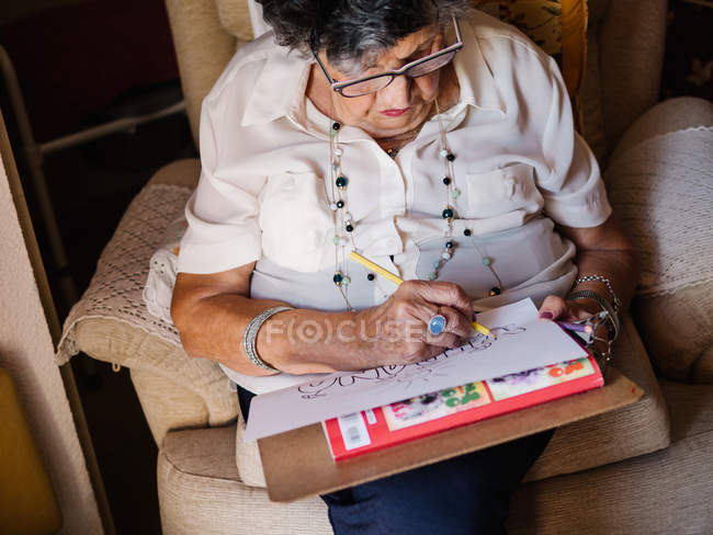 Mulher sênior em camisa branca e óculos desenhando em papel com lápis, sentado em poltrona em casa — Fotografia de Stock