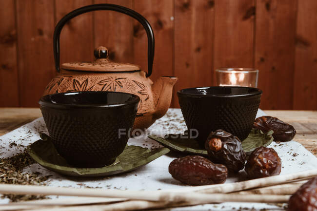 Сверху ароматный вкусный чай в чашке глиняный чайник и сладкие финики на белом подносе украшены чайными листьями на деревянном фоне — стоковое фото