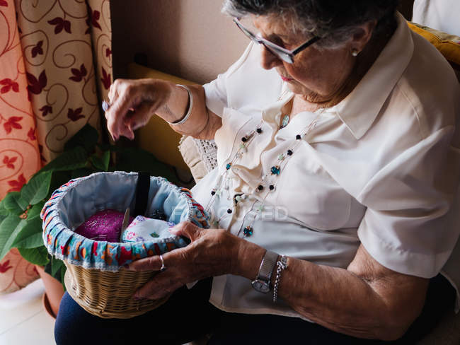 Von oben eine Seniorin in Bluse und Brille, die in der Wohnung Strickwerkzeuge aus Weidenkorb auswählt — Stockfoto