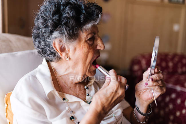 Mujer mayor que aplica lápiz labial mientras se mira en el espejo y se sienta en el sillón en casa - foto de stock