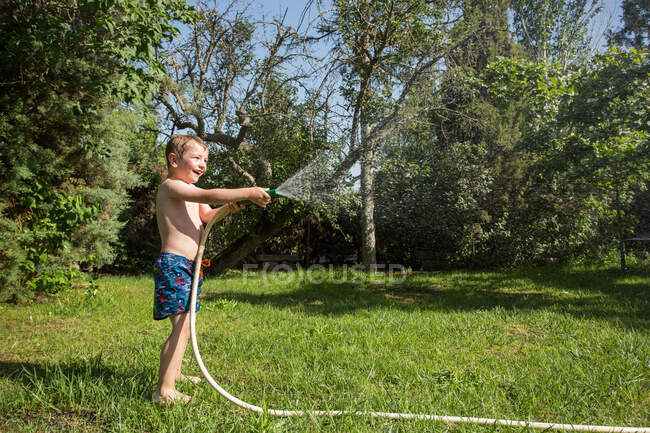 Pequeno garoto rindo em shorts e com os pés descalços salpicando água da mangueira de jardim — Fotografia de Stock