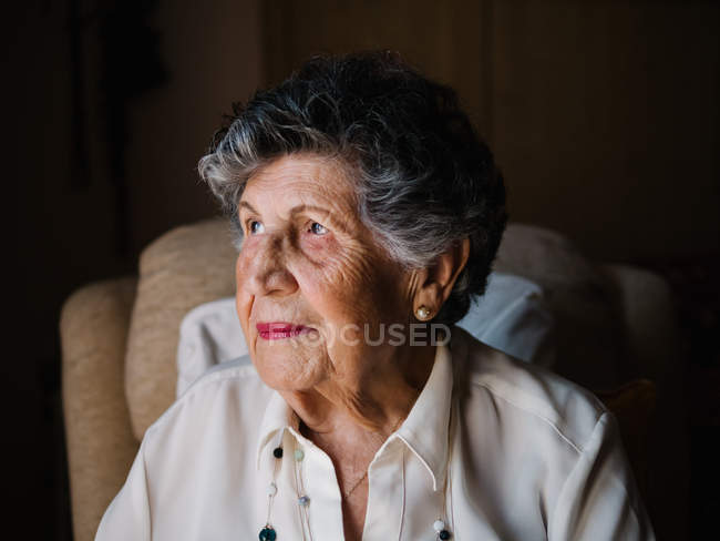 Портрет счастливой пожилой женщины в белой рубашке и с бусами на шее, смотрящей в камеру дома — стоковое фото