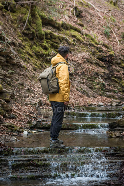Seitenansicht eines anonymen Mannes mit Rucksack, der auf natürlichen Stufen mit fließendem Wasser und in bergigem Gelände steht — Stockfoto