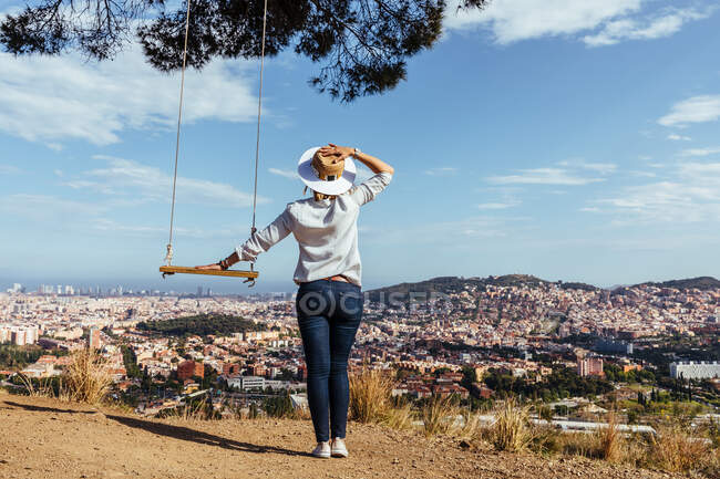 Vista de trás de uma menina com um chapéu olhando para a cidade ao lado de um balanço — Fotografia de Stock