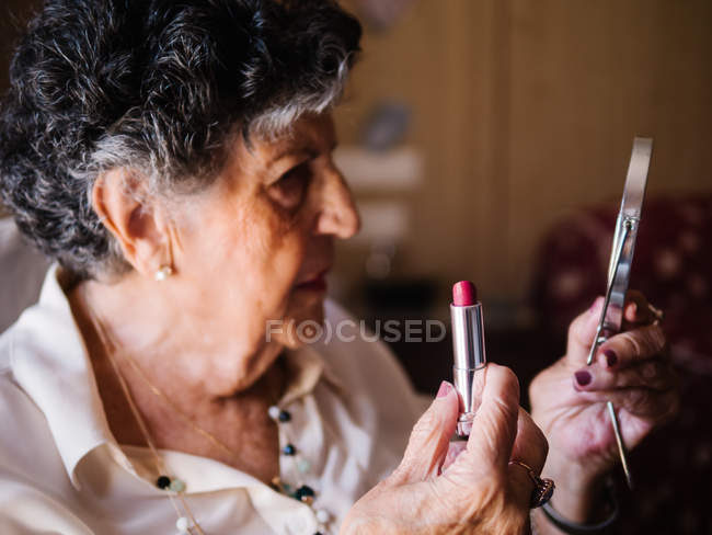 Mulher sênior aplicando batom enquanto olha no espelho e sentado na poltrona em casa — Fotografia de Stock