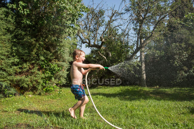 Kleines lachendes Kind in kurzen Hosen und mit nackten Füßen, die Wasser aus Gartenschlauch spritzen — Stockfoto