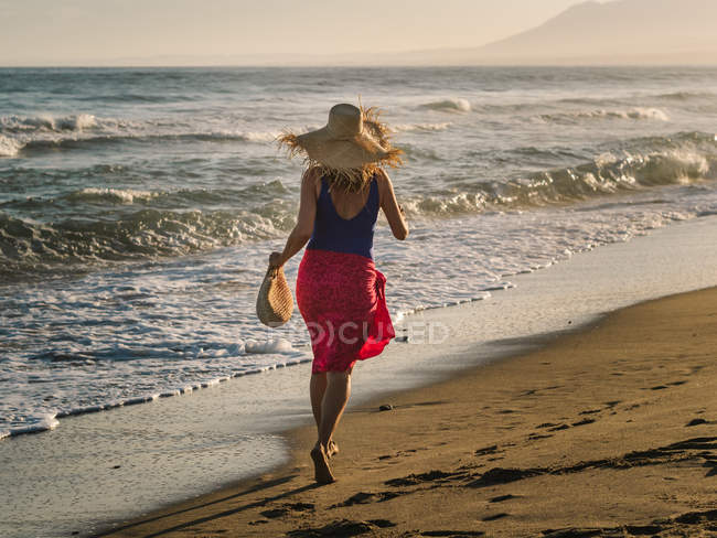 Вид ззаду струнка жінка в капелюсі і парео біжить уздовж берегової лінії в яскравому сонячному світлі — стокове фото