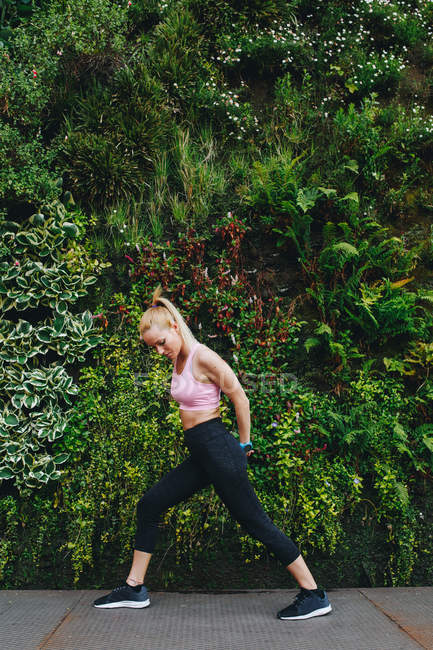 Mujer rubia joven haciendo ejercicios de alargamiento después de una sesión de ejecución con fondo de hierbas verdes - foto de stock