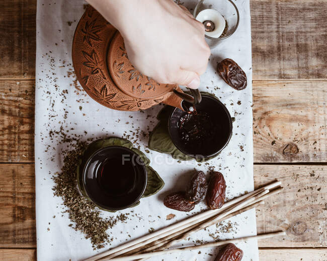 Mão pessoa irreconhecível servindo chá saboroso fragrante em xícara bule de barro e datas doces na bandeja branca decorada com folhas de chá no fundo de madeira — Fotografia de Stock