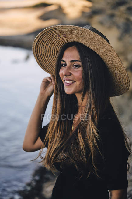 Молодая длинноволосая улыбающаяся женщина в шляпе смотрит вдаль на пляже — стоковое фото