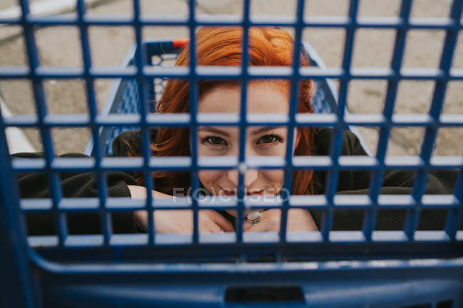 Attractive young woman with red hair sitting in blue shopping cart and looking in camera through trolley grate — Stock Photo