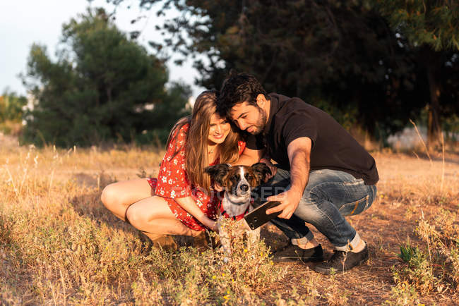 Sonriendo alegre pareja sentado entre la hierba alta y divertirse con poco perro en el campo - foto de stock