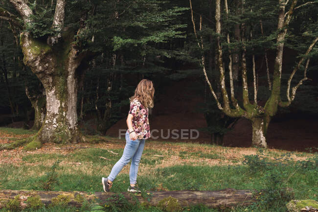 Seitenansicht einer jungen Touristin mit langen Haaren, die auf einem Baumstamm entlang des dunklen alten mystischen Waldes spaziert — Stockfoto