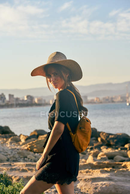 Young female tourist wearing a straw hat and backpack standing near beach — Stock Photo