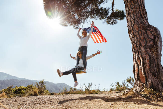 Two young girls with an American flag celebrating the 4th of July on a swing — Stock Photo