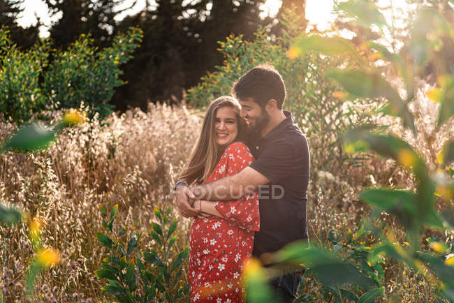 Людина обіймає усміхнену вагітну дружину на фоні мальовничого зеленого парку в сонячний день — стокове фото