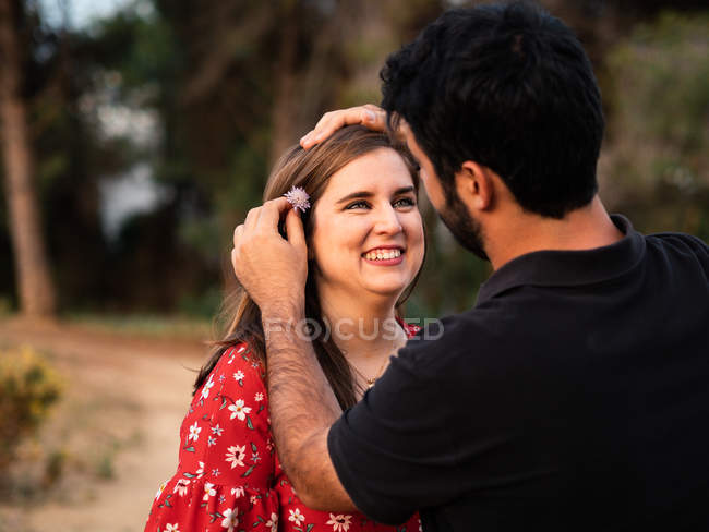 L'uomo sorridente a moglie in natura sfocata e mettendo il fiore in capelli — Foto stock