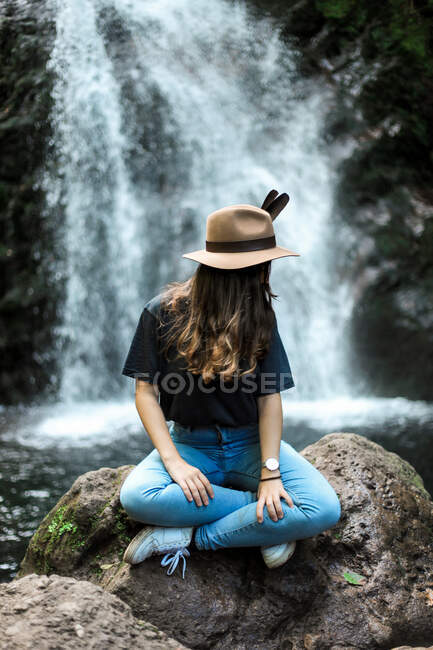 Allegro viaggiatore femminile in cappello sorridente e guardando la fotocamera mentre seduto sul masso bagnato vicino alla cascata — Foto stock