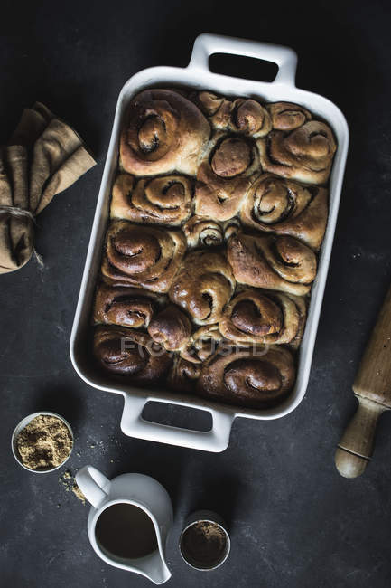 From above tray with fresh homemade cinnamon buns and wooden rolling pin on black background — Stock Photo