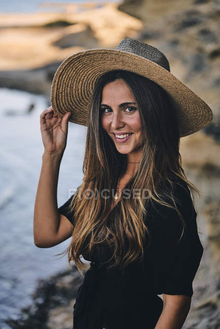 Joven mujer de pelo largo sonriente en sombrero mirando a la cámara en la playa - foto de stock