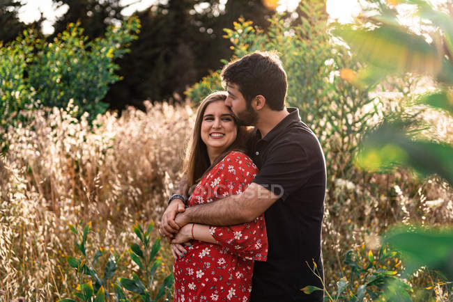 Hombre pensativo abrazando sonriente esposa embarazada en el fondo del pintoresco parque verde en el día soleado - foto de stock