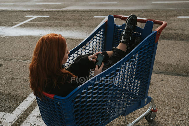 Woman with smartphone in shopping trolley in parking lot — Stock Photo