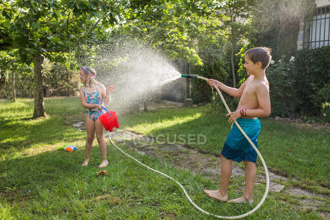 Ragazzo in costume da bagno spruzzando acqua dal tubo da giardino a una ragazza in una giornata di sole — Foto stock