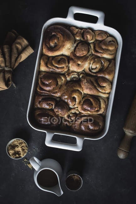 From above tray with fresh homemade cinnamon buns and wooden rolling pin on black background — Stock Photo