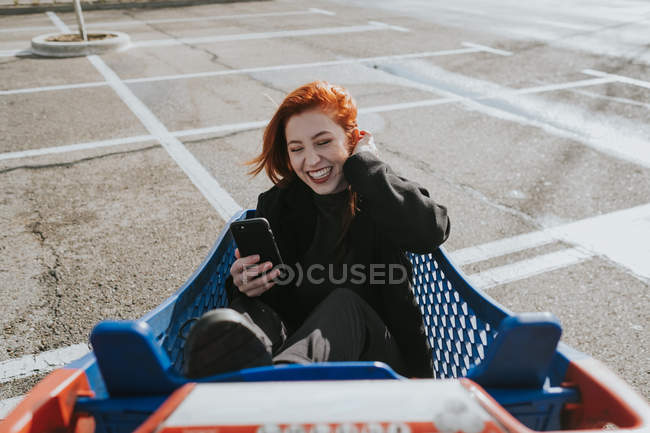 Smiling woman with smartphone in shopping trolley in parking lot — Stock Photo