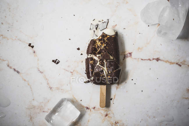 Ghiacciolo di gelato alla vaniglia e cioccolato schiantato sulla superficie di marmo con cubetti di ghiaccio — Foto stock