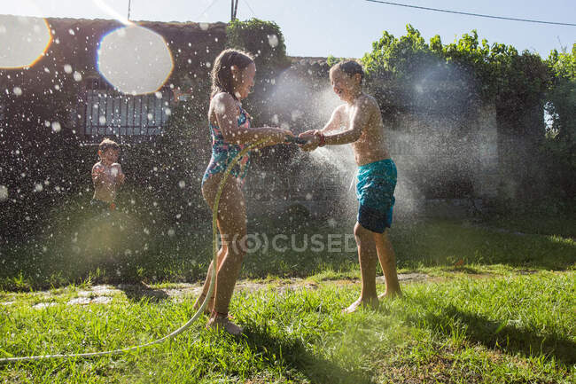 Niños pequeños en traje de baño corriendo y salpicando agua de la manguera del jardín entre sí - foto de stock