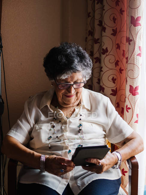 Anziana in camicia e occhiali seduta su poltrona e lettura e-book in appartamento — Foto stock