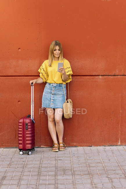 Young female tourist using mobile phone while leaning on red wall — Stock Photo