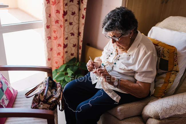 Mulher sênior em blusa e óculos escolhendo ferramentas de tricô de cesta de vime, sentado em apartamento — Fotografia de Stock