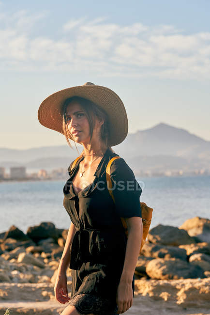 Молода жінка-туристка в солом'яному капелюсі і рюкзаку, що стоїть біля пляжу — стокове фото