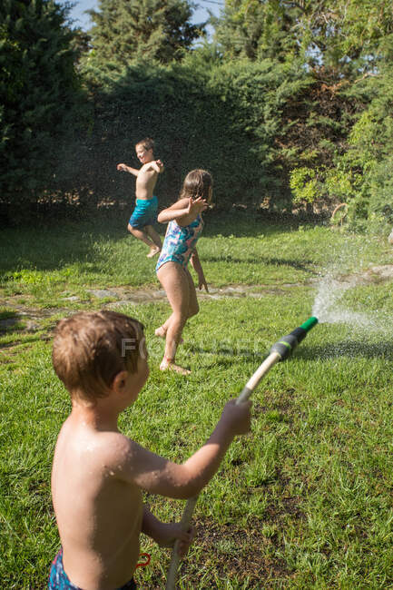 Kleine Kinder in Badebekleidung rennen herum und spritzen sich gegenseitig Wasser aus Gartenschlauch zu — Stockfoto