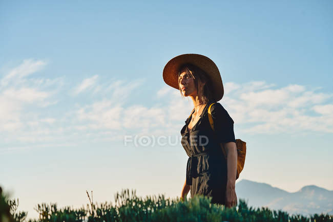 Turista femenina con sombrero de paja y mochila de pie en la naturaleza - foto de stock