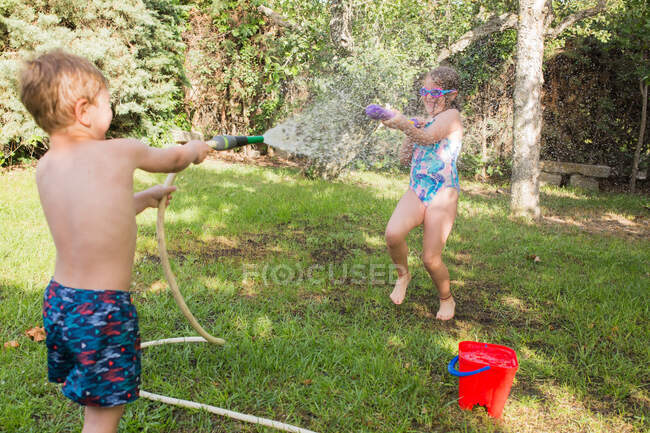 Niños pequeños en traje de baño corriendo y salpicando agua de la manguera del jardín entre sí - foto de stock