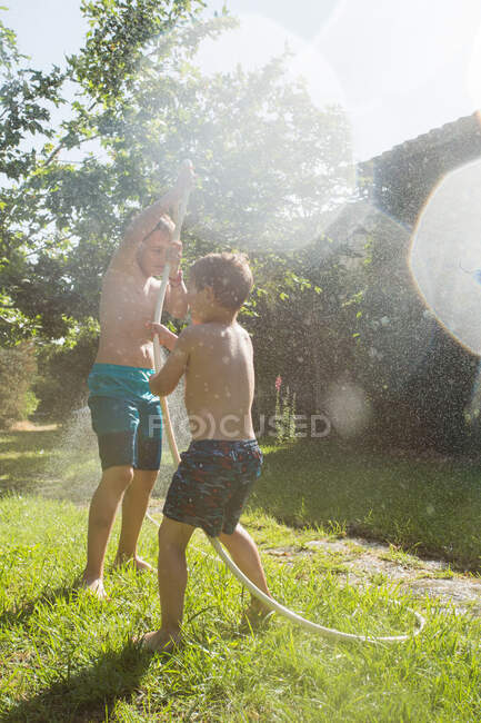 I bambini piccoli in costume da bagno che corrono in giro e spruzzano acqua dal tubo da giardino l'uno contro l'altro — Foto stock