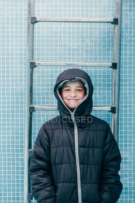 Niño sonriente en chaqueta caliente de pie sobre el fondo de la pared de la piscina y mirando a la cámara - foto de stock