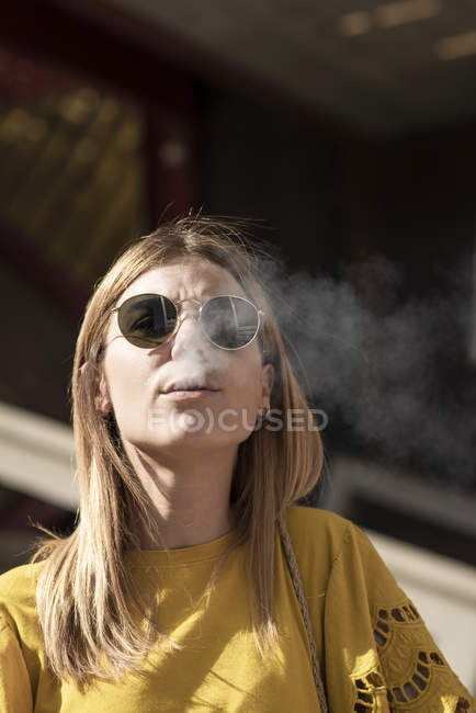Giovane donna elegante che indossa occhiali da sole soffiando fumo di sigaretta — Foto stock