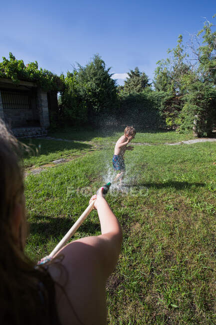 Niños pequeños en traje de baño corriendo y salpicando agua de la manguera del jardín entre sí, vista en primera persona - foto de stock