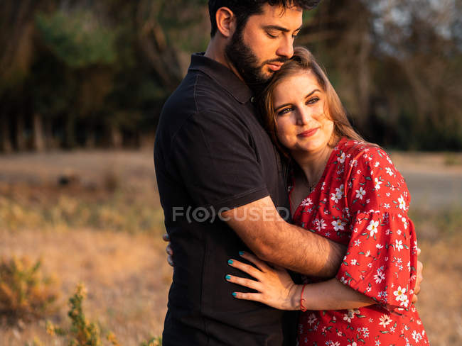 Чоловік обіймає усміхнену вагітну дружину на фоні природи в сонячний день — стокове фото