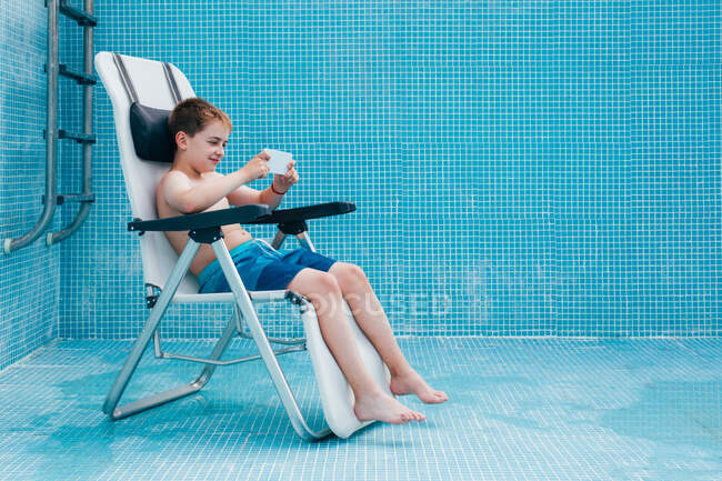 Menino com smartphone sentado no fundo da piscina vazia — Fotografia de Stock