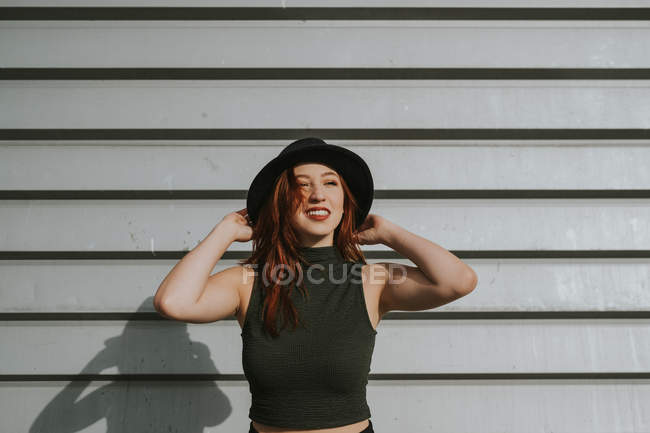 Atractiva joven con el pelo rojo en sombrero negro sosteniendo sombrero y sonriendo contra la pared áspera - foto de stock